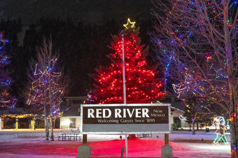 Nighttime view of the Red River, New Mexico sign with welcoming message, surrounded by trees decorated with colorful lights and a prominent, large red tree with a star on top, illuminated against a dark sky.