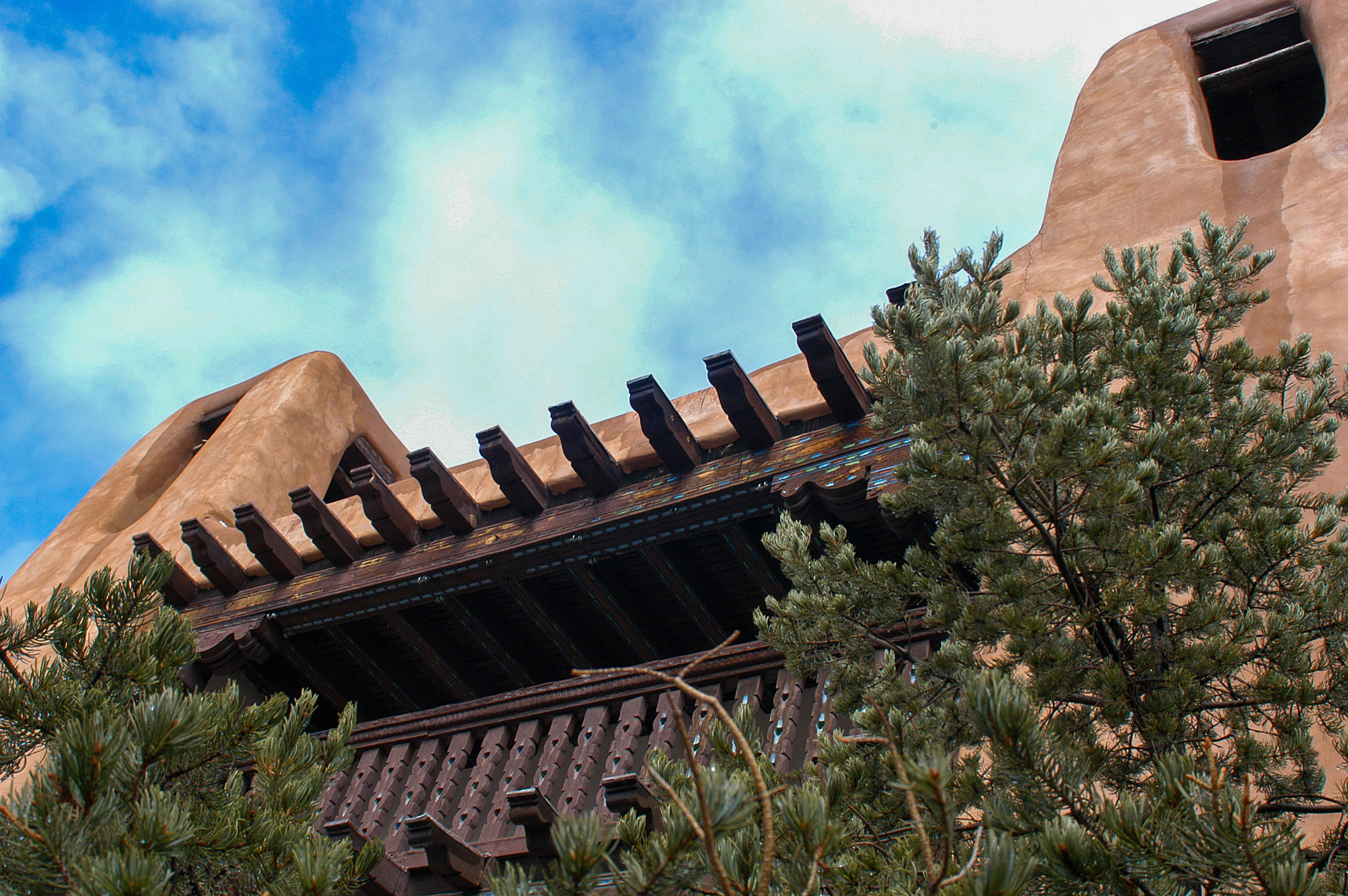Adobe-style building with distinctive wooden beams under a vibrant sky, surrounded by pine trees.