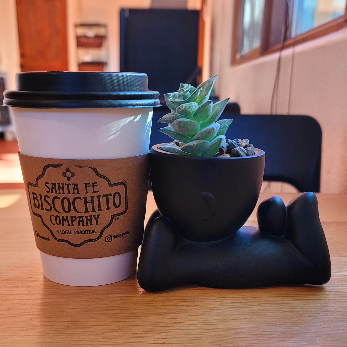 A paper coffee cup with "Santa Fe Biscochito Company" logo next to a small succulent plant in a quirky pot shaped like a reclining figure, placed on a wooden table.