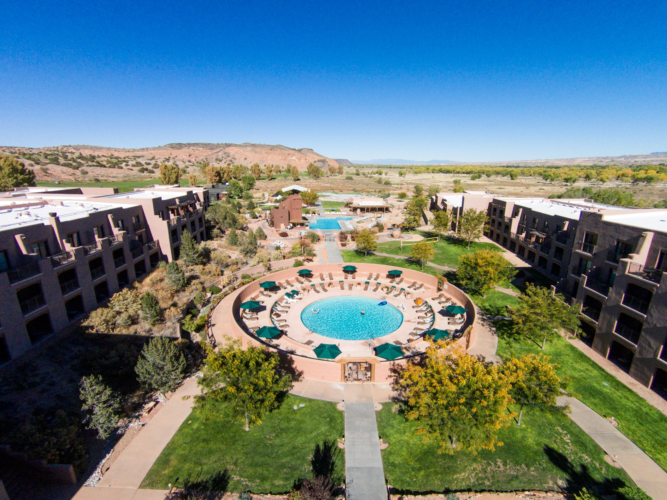 Aerial view of a resort with a circular pool, landscaped gardens, and surrounding buildings under a clear blue sky.
