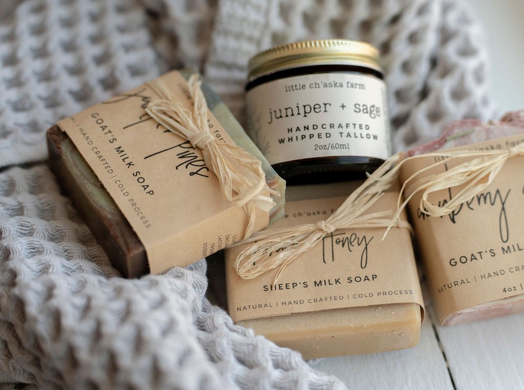 Handcrafted soaps and salves labeled "Oat & Honey" and "Juniper & Sage" from Little Chaska Farm, arranged on a soft knitted grey blanket.