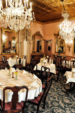 An elegant dining room featuring tables set with white table cloths and flowers, sitting beneath ornate chandeliers