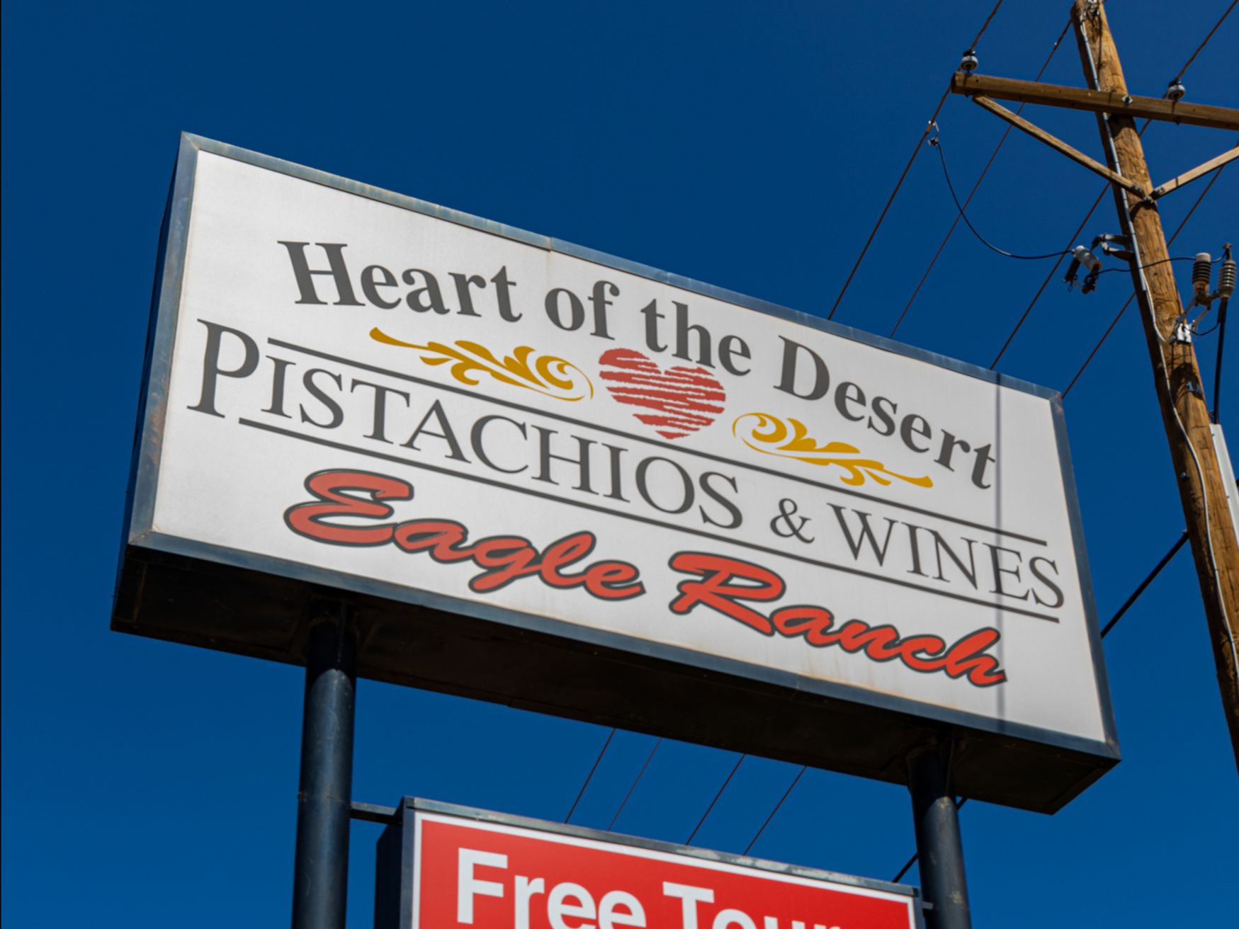Signboard for "Heart of the Desert Pistachios & Wines at Eagle Ranch," advertising free tours, a gift shop, and free wine tasting under a clear blue sky.