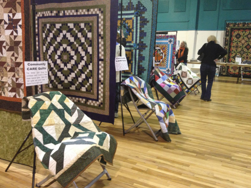 Quilt display at a community event in a large hall, featuring intricate patterns and various designs on both standing frames and draped over chairs. People are examining the quilts.