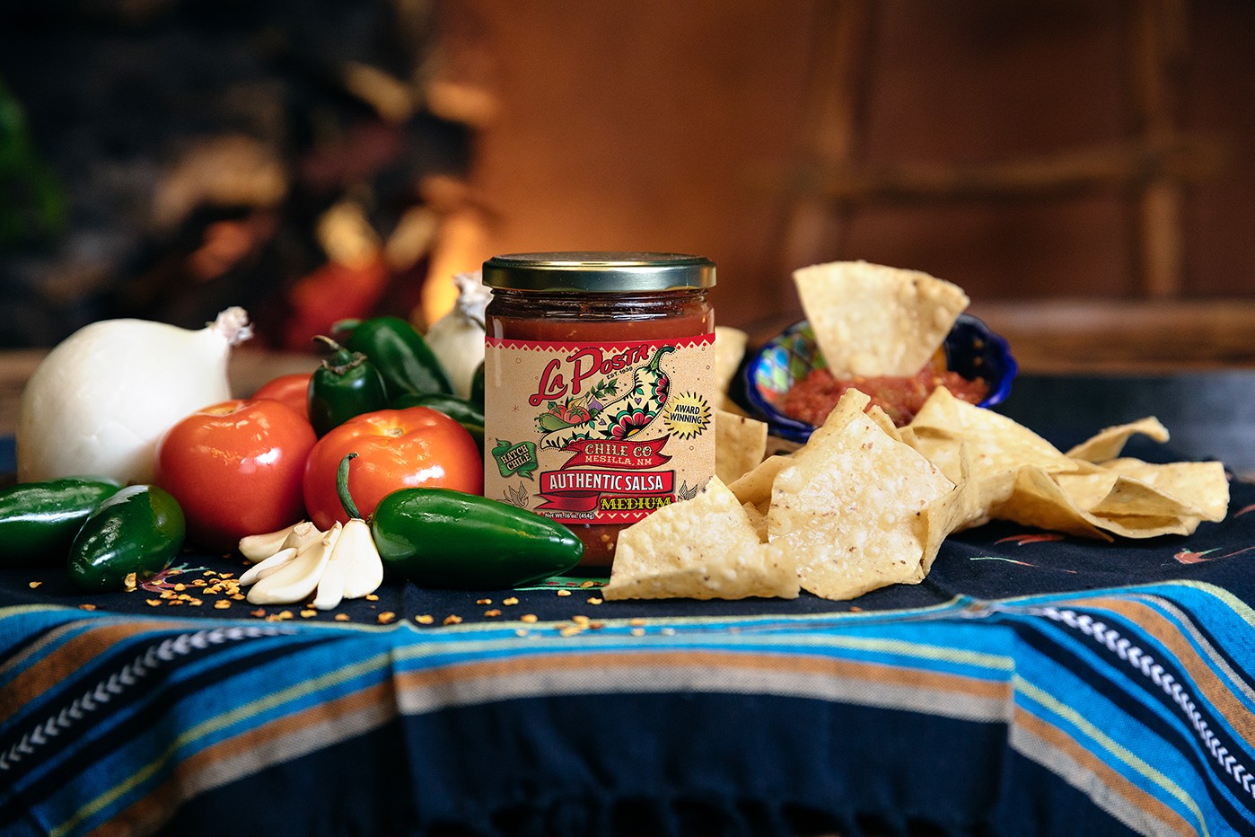 A jar of La Posta's Authentic Salsa surrounded by tortilla chips, jalapeños, tomatoes, onions, and chips on a colorful striped tablecloth.