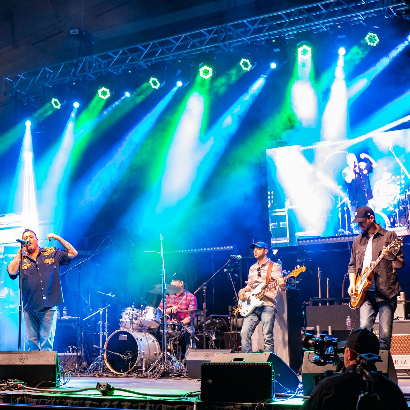 A rock band performs live on stage under vibrant green and blue stage lighting, with a dynamic display screen in the background enhancing the concert atmosphere.
