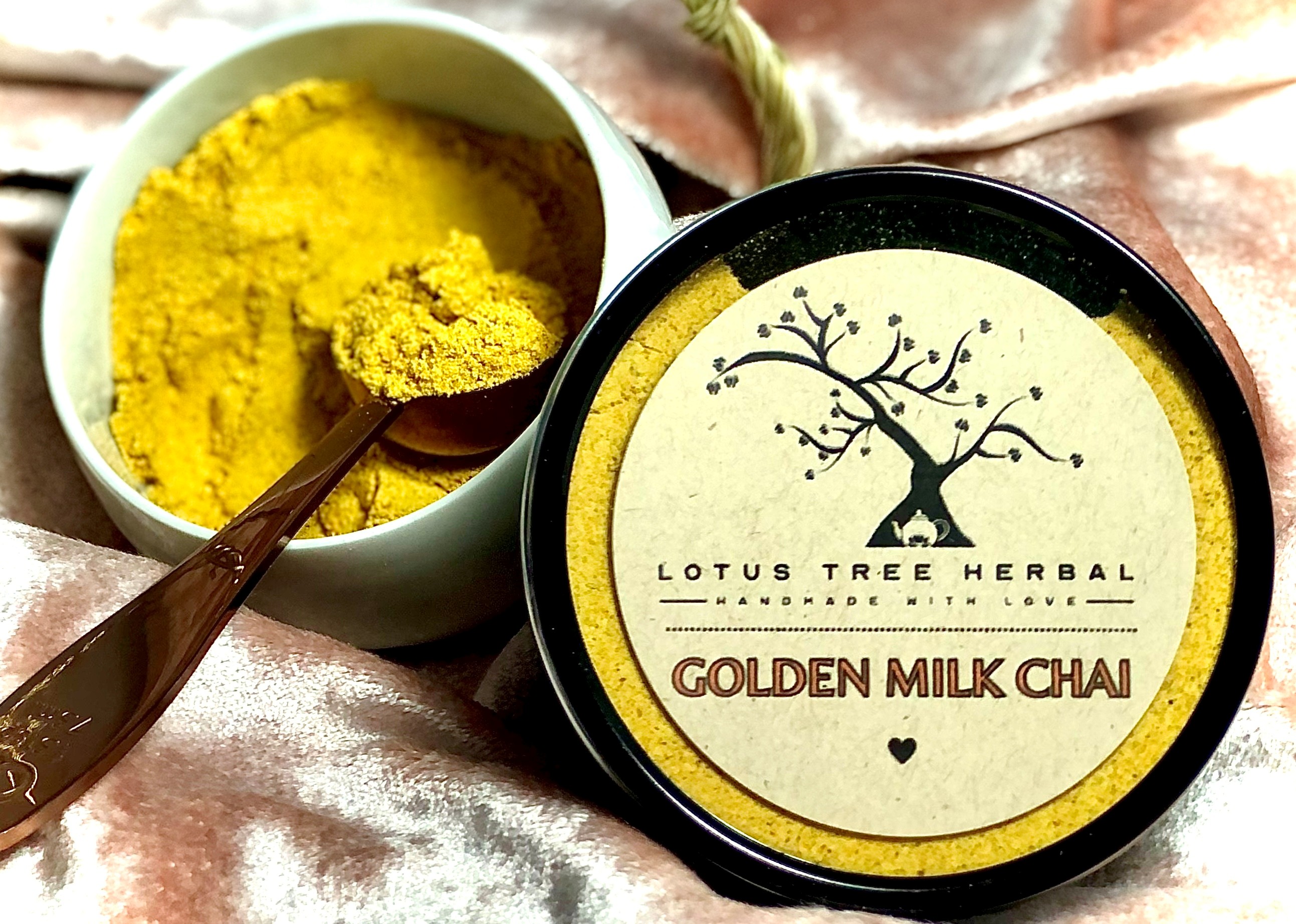 A container of Lotus Tree Herbal Golden Milk Chai next to a bowl filled with yellow powder and a spoon on a textured pink surface.
