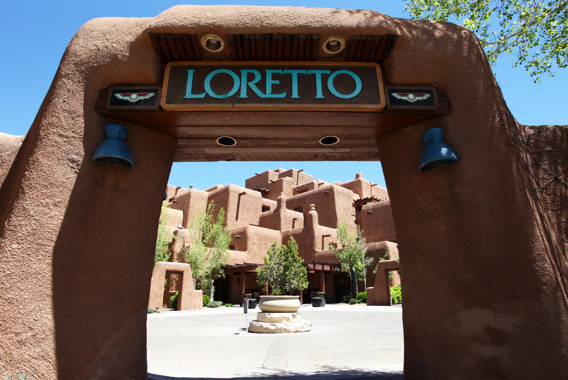 Entrance to the Loretto, featuring an adobe archway with the name "LORETTO" at the top and two blue bells, overlooking a courtyard with Southwestern-style architecture.