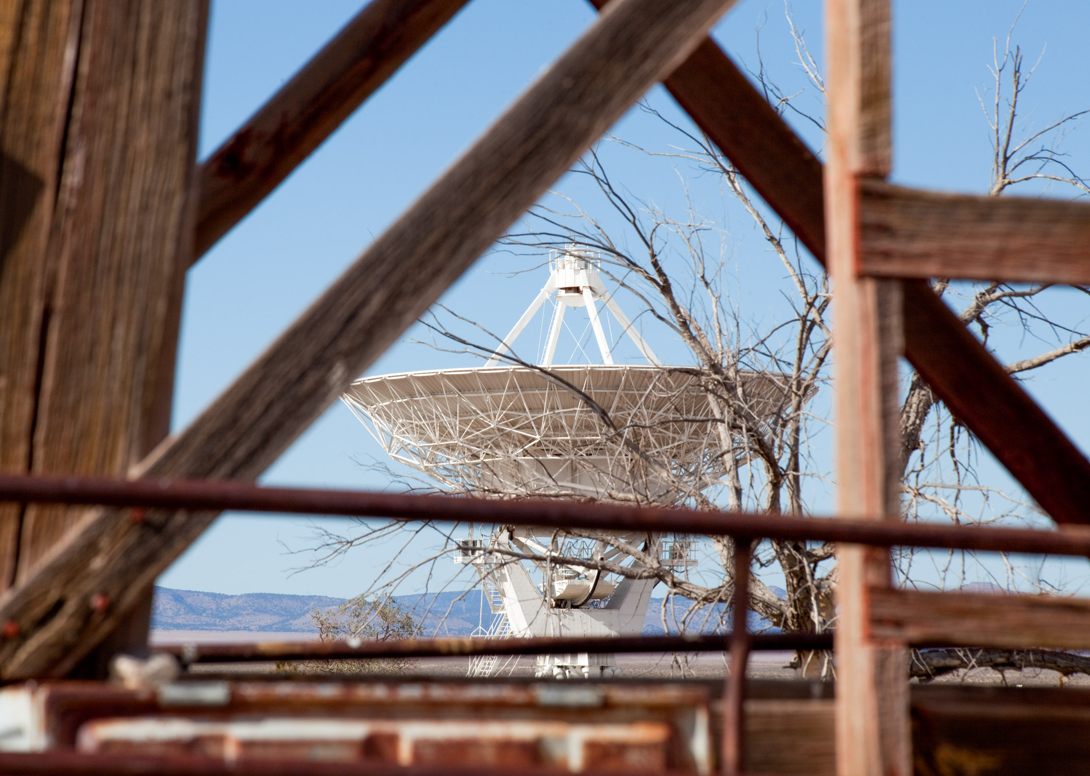 A large satellite dish viewed through a rustic wooden frame against a clear blue sky.