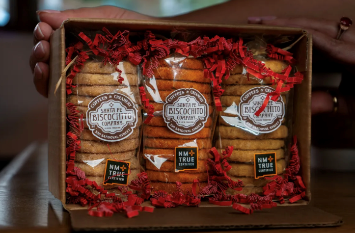 A person holding a wooden gift box filled with packages of Santa Fe Biscochito Company cookies. The packaging features prominent labels and is surrounded by decorative red filler.
