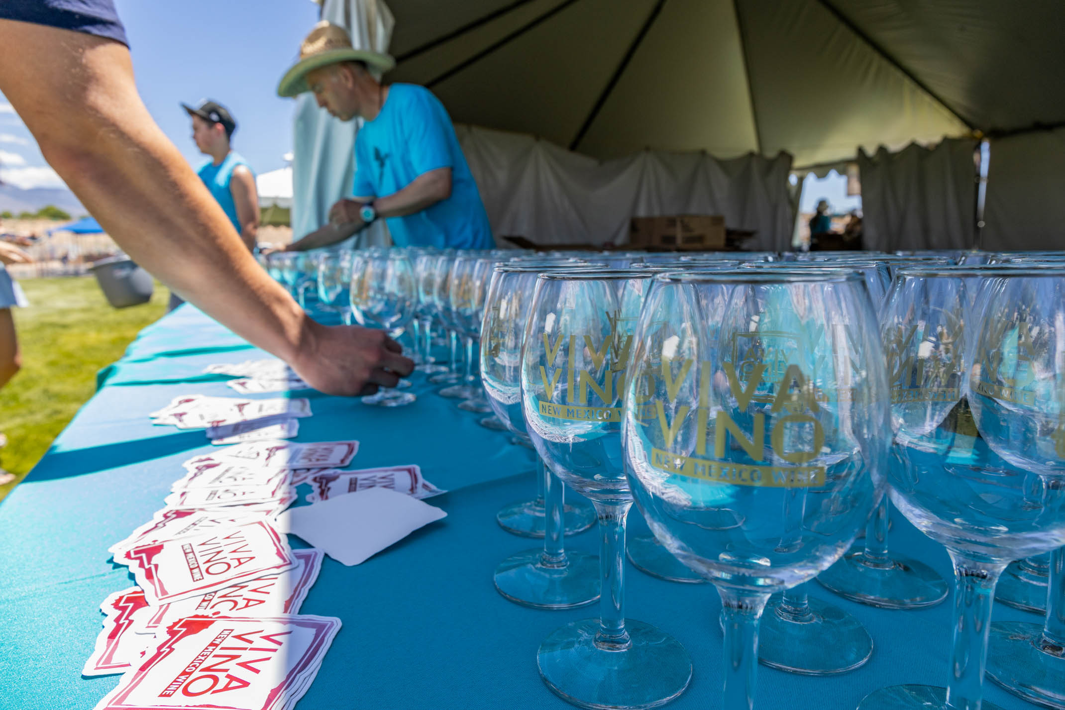 A person reaches for a one wine glass out of a table-full under a tent, featuring stickers sitting on the table.