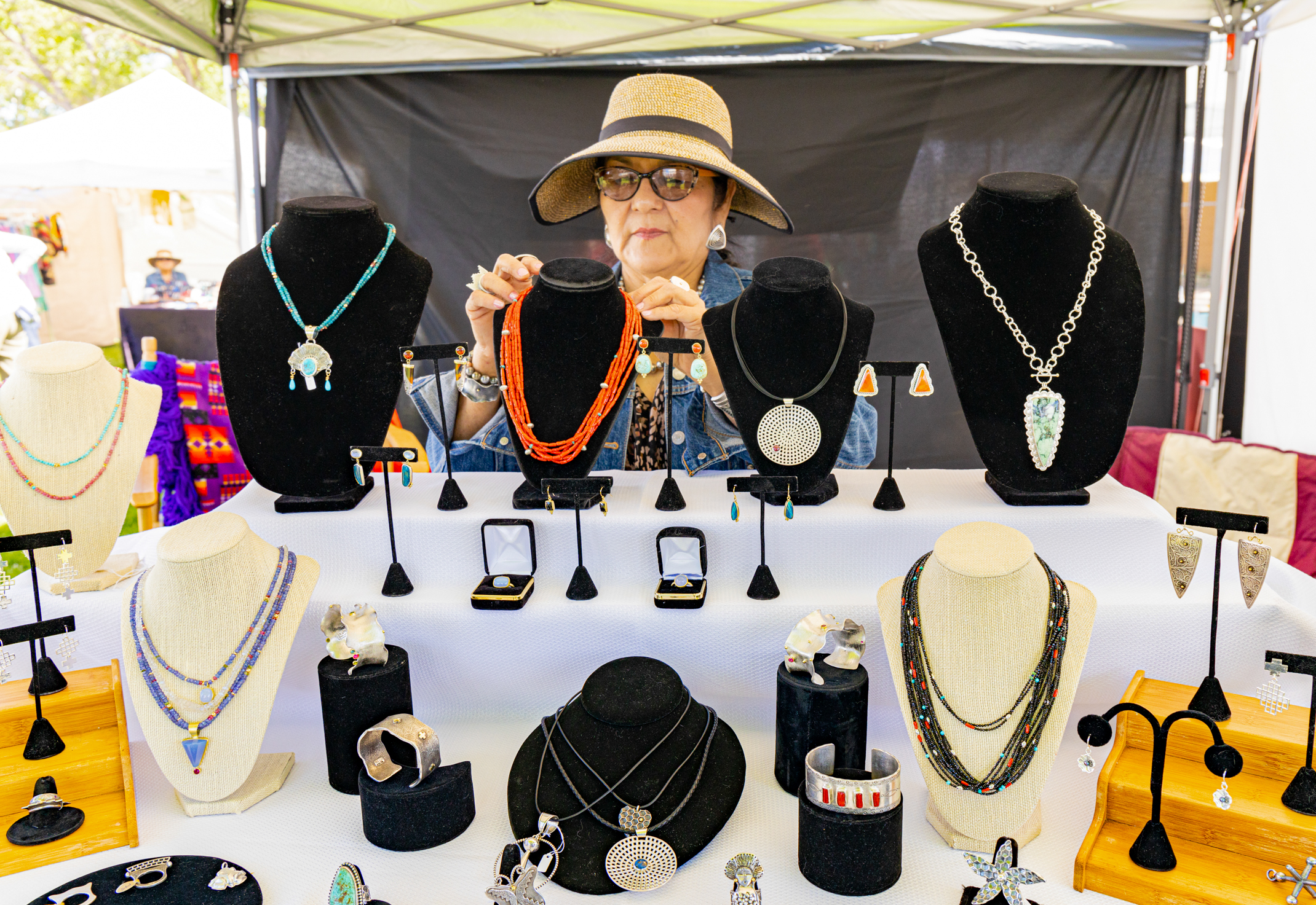 A person wearing a wide-brimmed hat and sunglasses sits behind a table displaying various pieces of jewelry at an outdoor market.