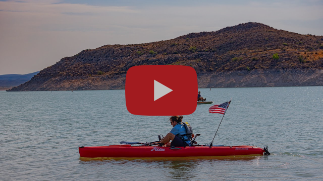 A person sits in a red kayak floating in a lake with a large hill in the background.