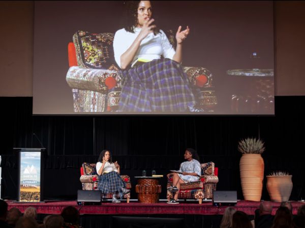Two people sitting on a stage in chairs with a screen behind them broadcasting them for the crowd.