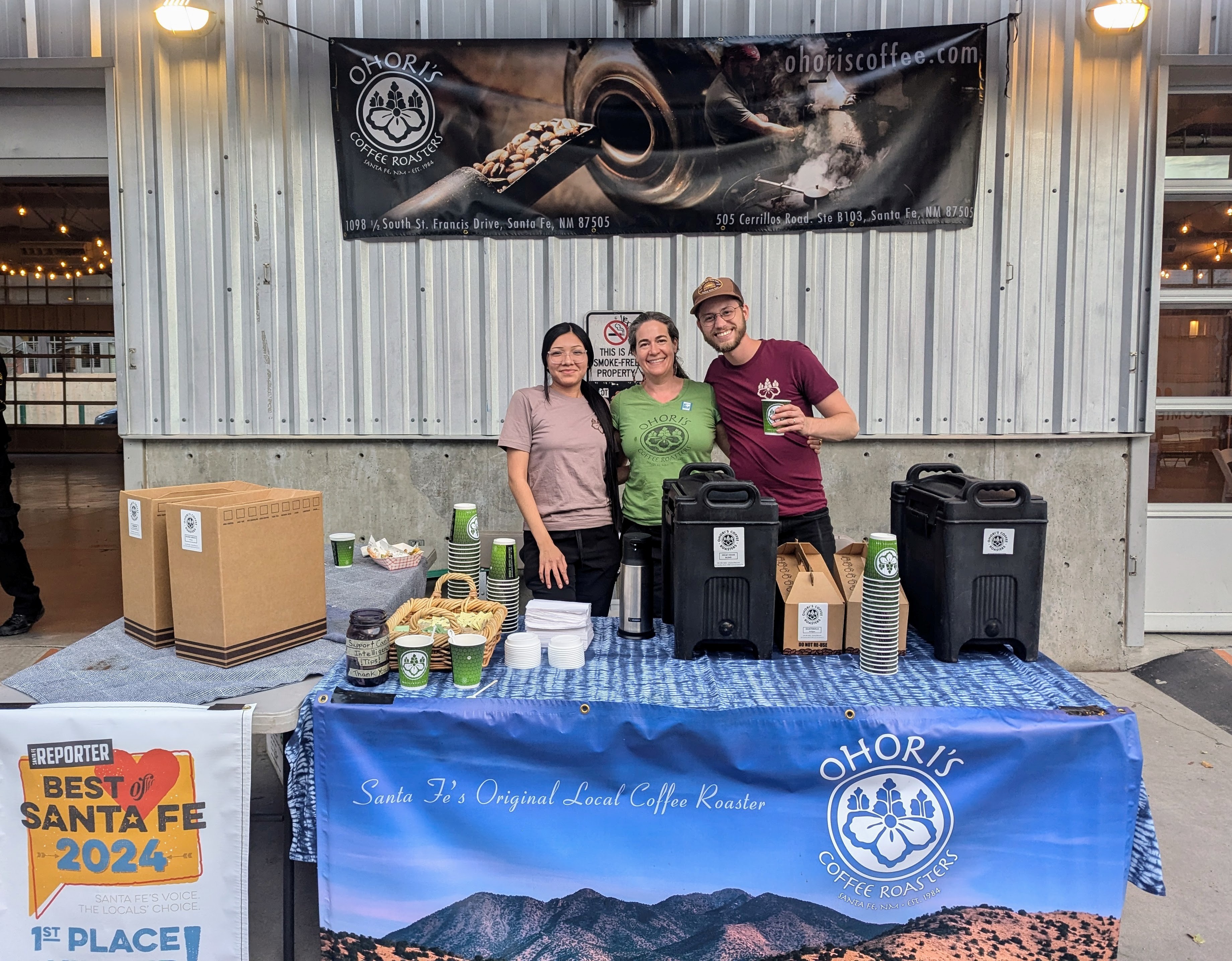 Three people standing behind a table with different receptacles and coffee cups on top of it. The banner on the table reads Ohori's Coffee Roasters