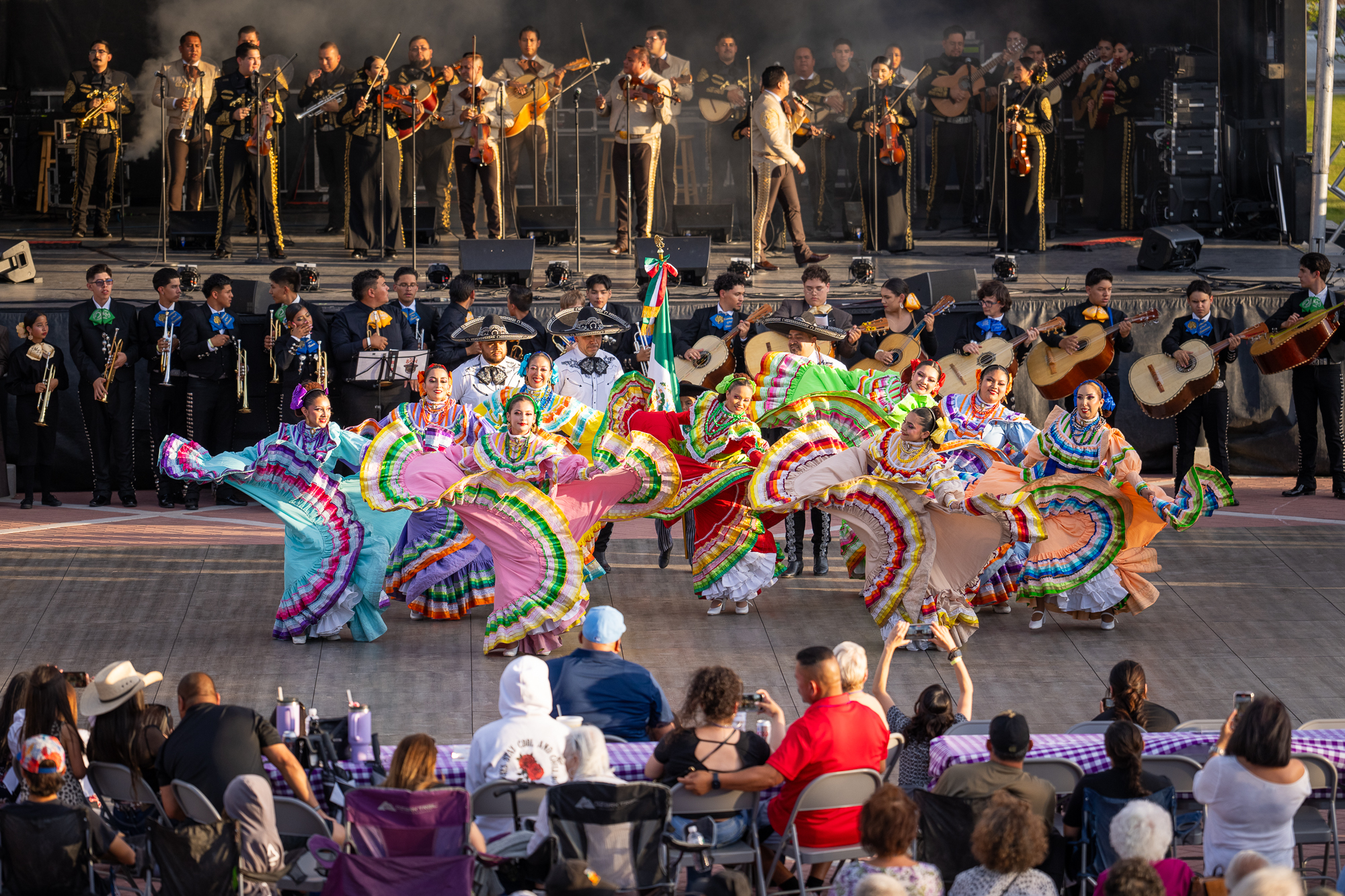 Performers in colorful traditional Mexican dresses dance in front of a large mariachi band on an outdoor stage, witnessed by an audience in bright daylight.