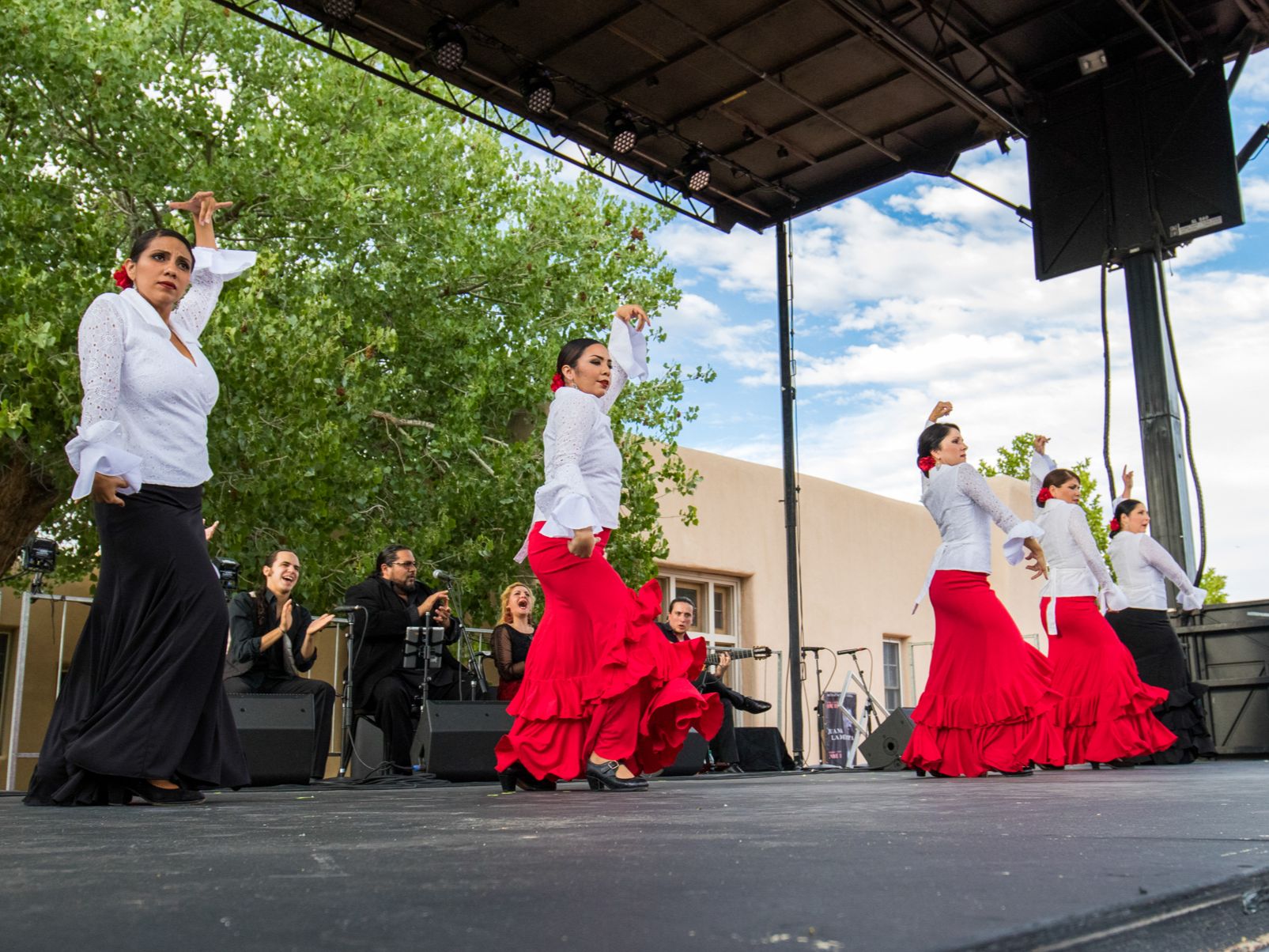 Performers in traditional red and white flamenco attire dancing on an outdoor stage, with musicians in black playing in the background.