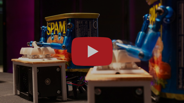Two cans of spam, modified in the form of humanoid robots, stand in front of a set of keys built into small tables