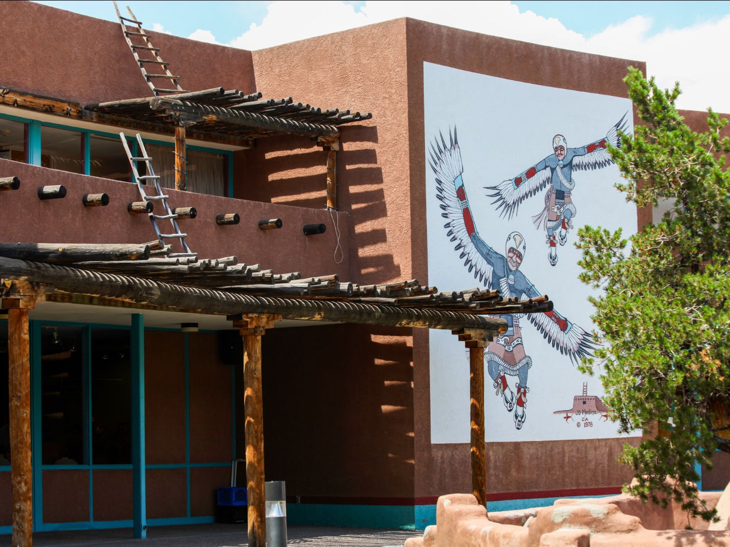 Mural of Indigenous Dancers in traditional garb depicted on the wall of a pueblo-style building with wooden vigas and turquoise door.