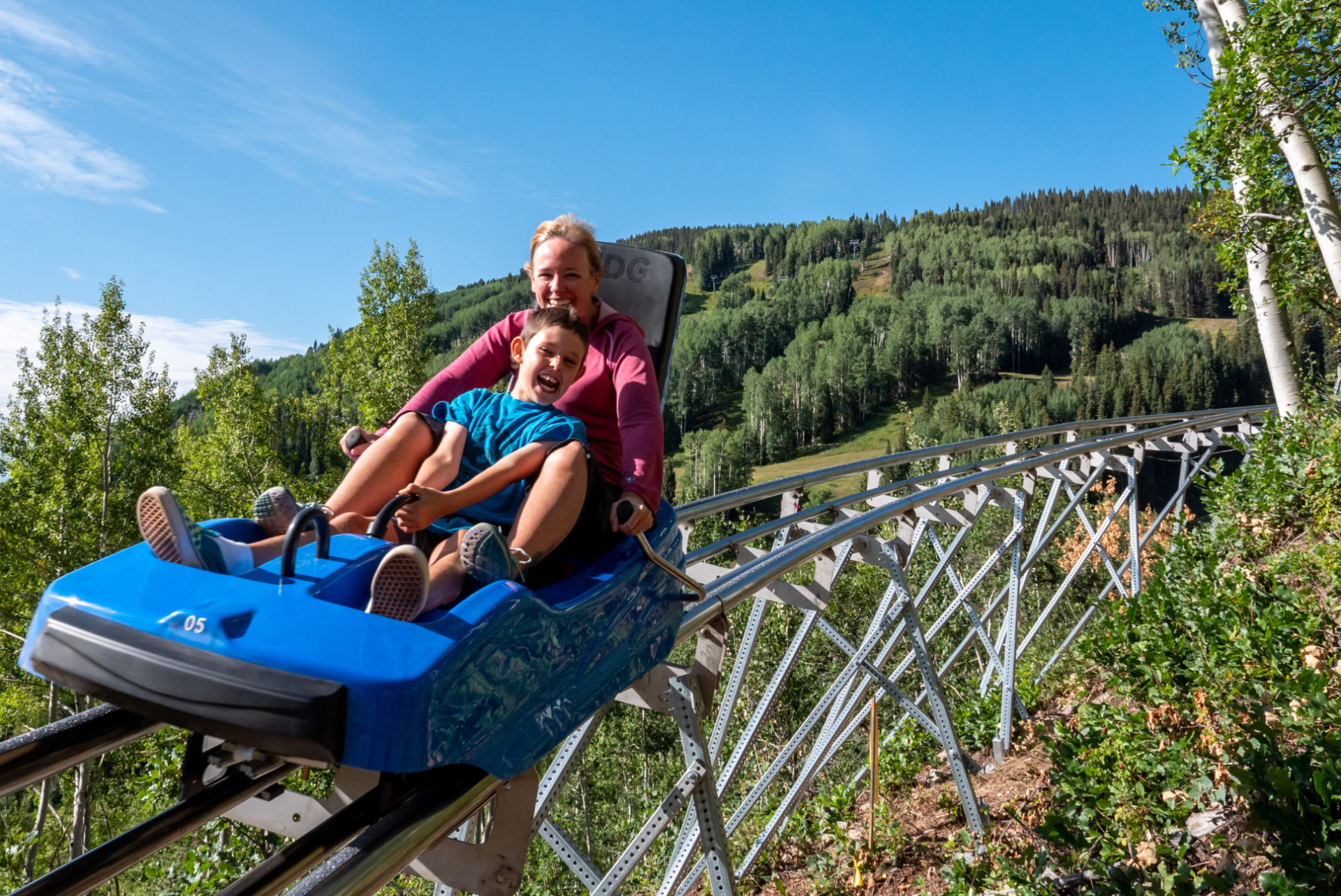 Two individuals smiling and riding a blue mountain coaster in a lush, green environment.