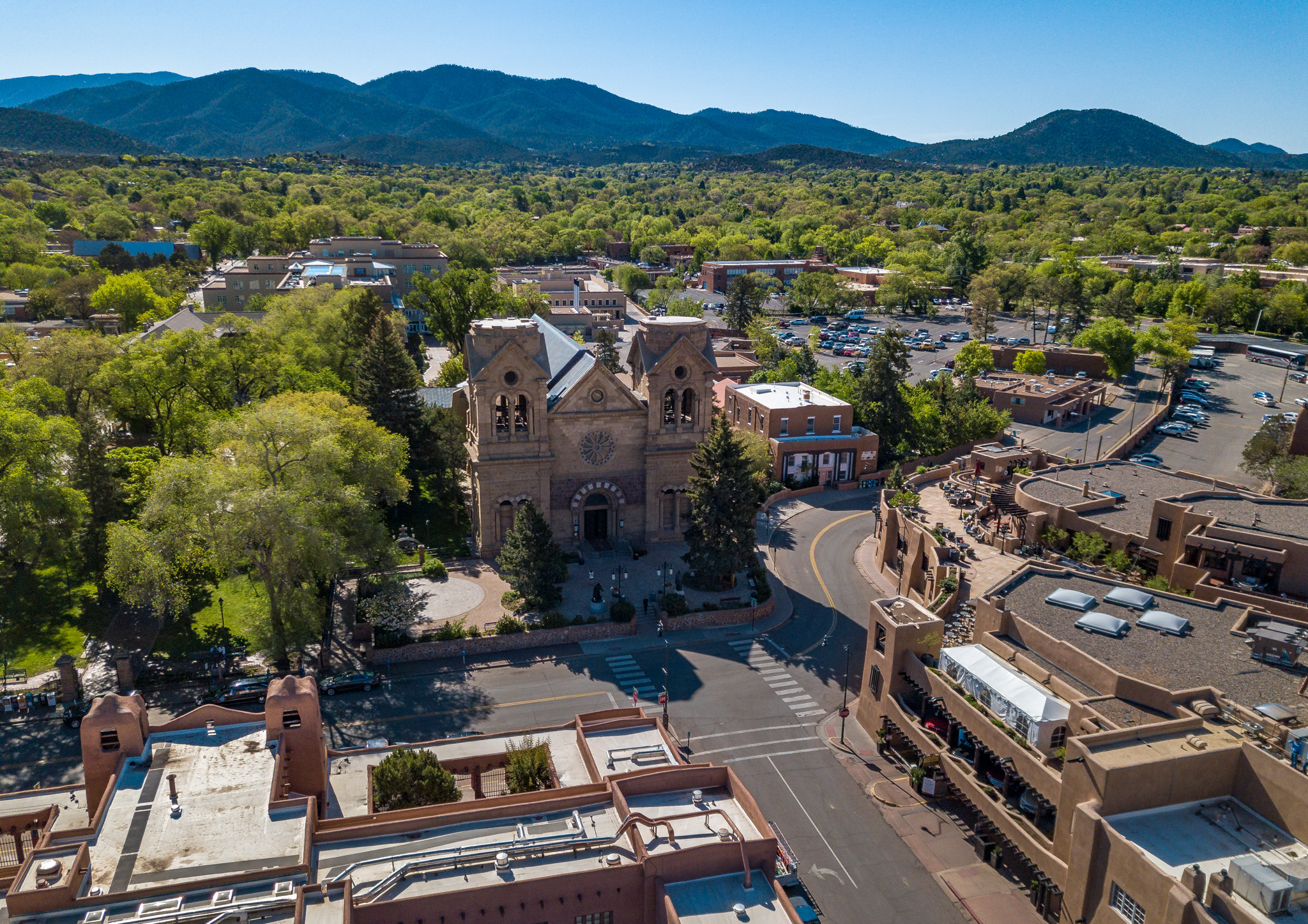 Aerial view of a large stone church surrounded by trees and other buildings and green trees, with mountains in the background.