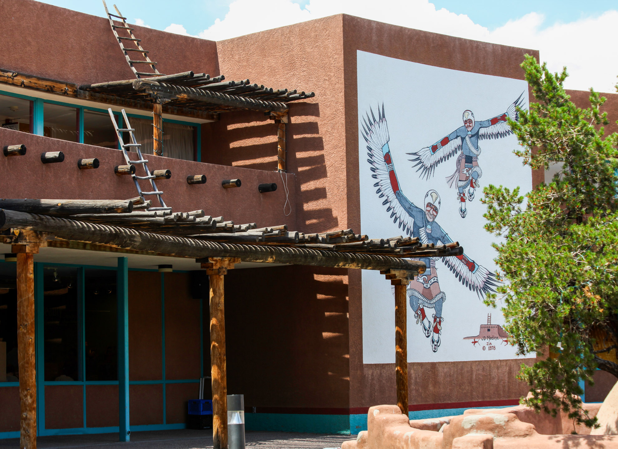 An adobe structure with a wooden log overhang that has a mural of Indigenous Dancers in traditional attire on the side