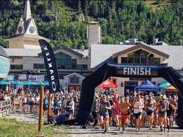 Runners crossing the finish line at a race event, with spectators cheering near a finish arch and a distinctive building with a pointed roof in the background.
