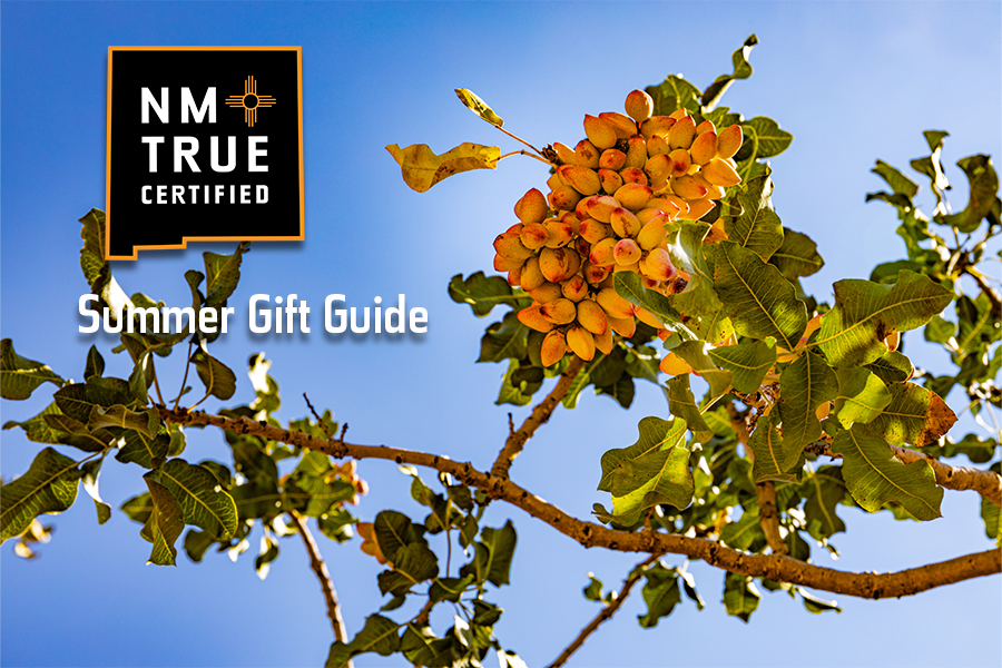 A bunch of ripening pistachios hang on a tree branch in front of a bright blue sky. A logo for NM True Certified is overlaid and text reads Summer Gift Guide