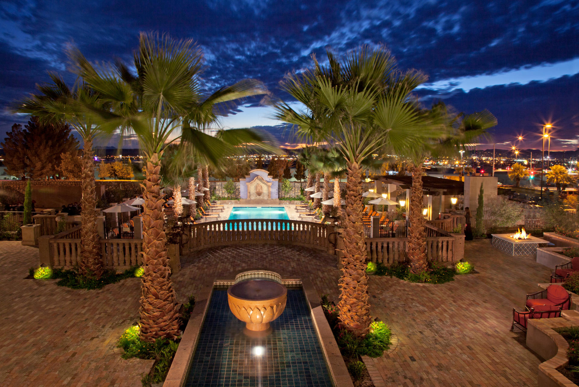 Nighttime view of an elegant outdoor swimming pool area surrounded by palm trees, with a scenic city skyline in the background.