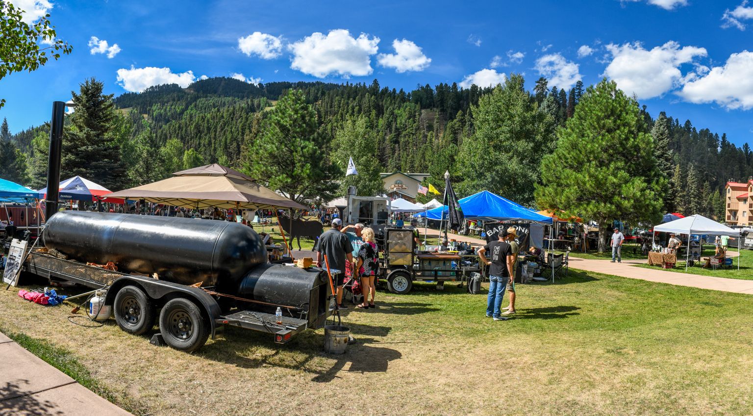 Panoramic view of an outdoor market in a grassy park with various vendor tents, people browsing, and a large barbecue smoker trailer under a clear blue sky.
