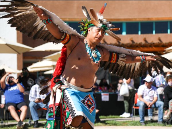 A person performs a traditional Native American dance in full regalia, featuring expansive feathered wings and intricate beadwork, at an outdoor cultural festival.