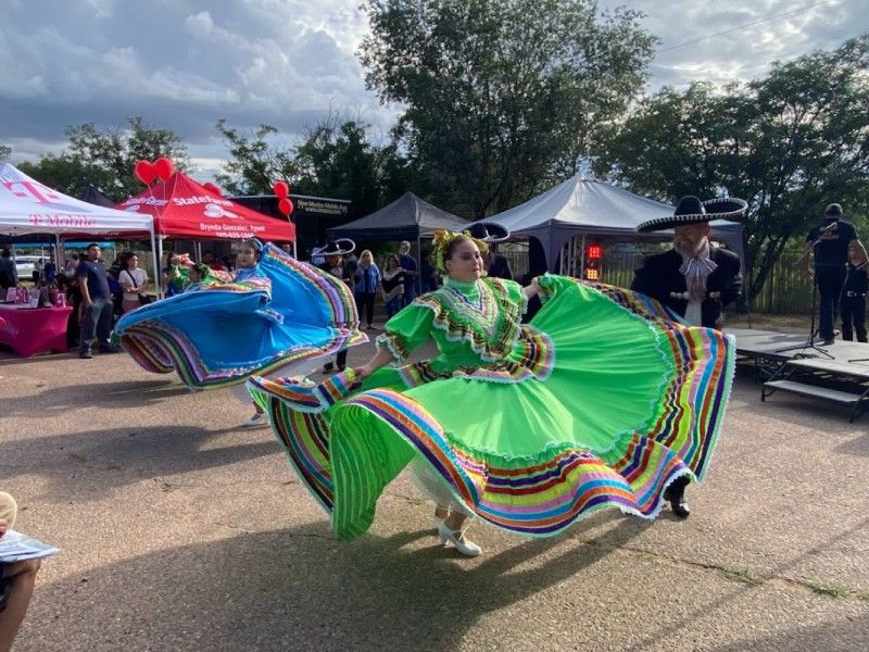 Performers in vibrant, multicolored traditional dresses dancing at an outdoor event, with spectators and tents in the background.