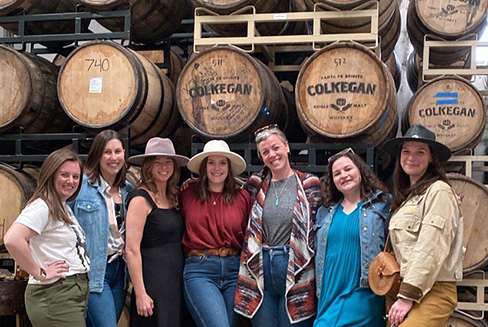 Group of seven individuals posing in front of Colkegan whiskey barrels, smiling.