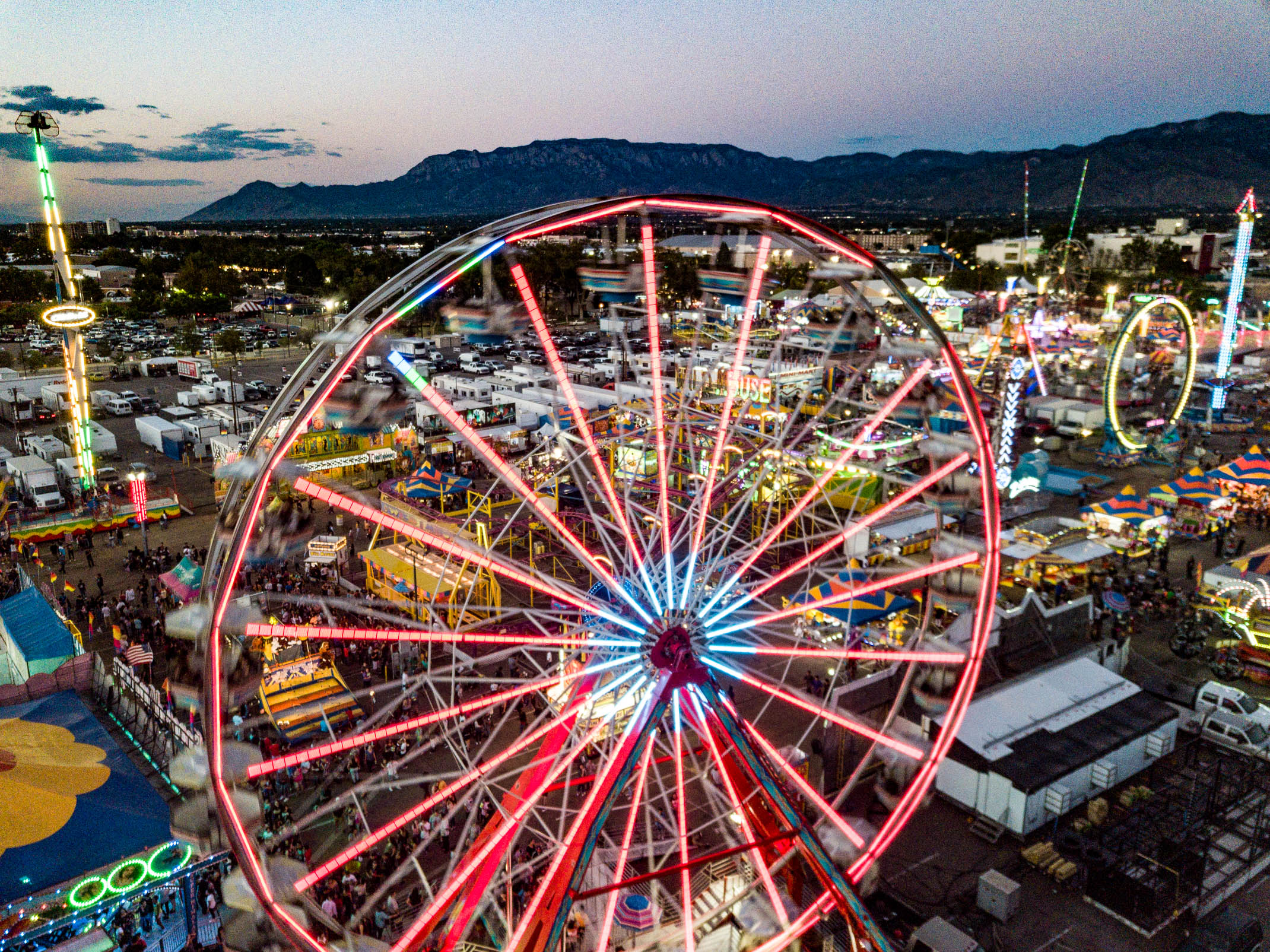 A large Ferris wheel covered in colorful LED's towers over other carnival rides and booths with silhouetted mountains in the distance