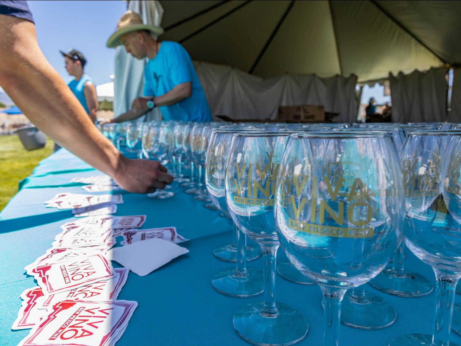 Wine glasses sit on a table as someone inspects them, text on the glasses reads Viva Vino
