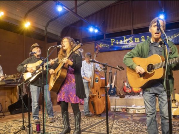 Musicians perform on stage at Passim, a music venue, with three guitar players in the foreground energetically playing and singing into microphones, accompanied by a violinist and a bass player in the background under warm stage lighting.