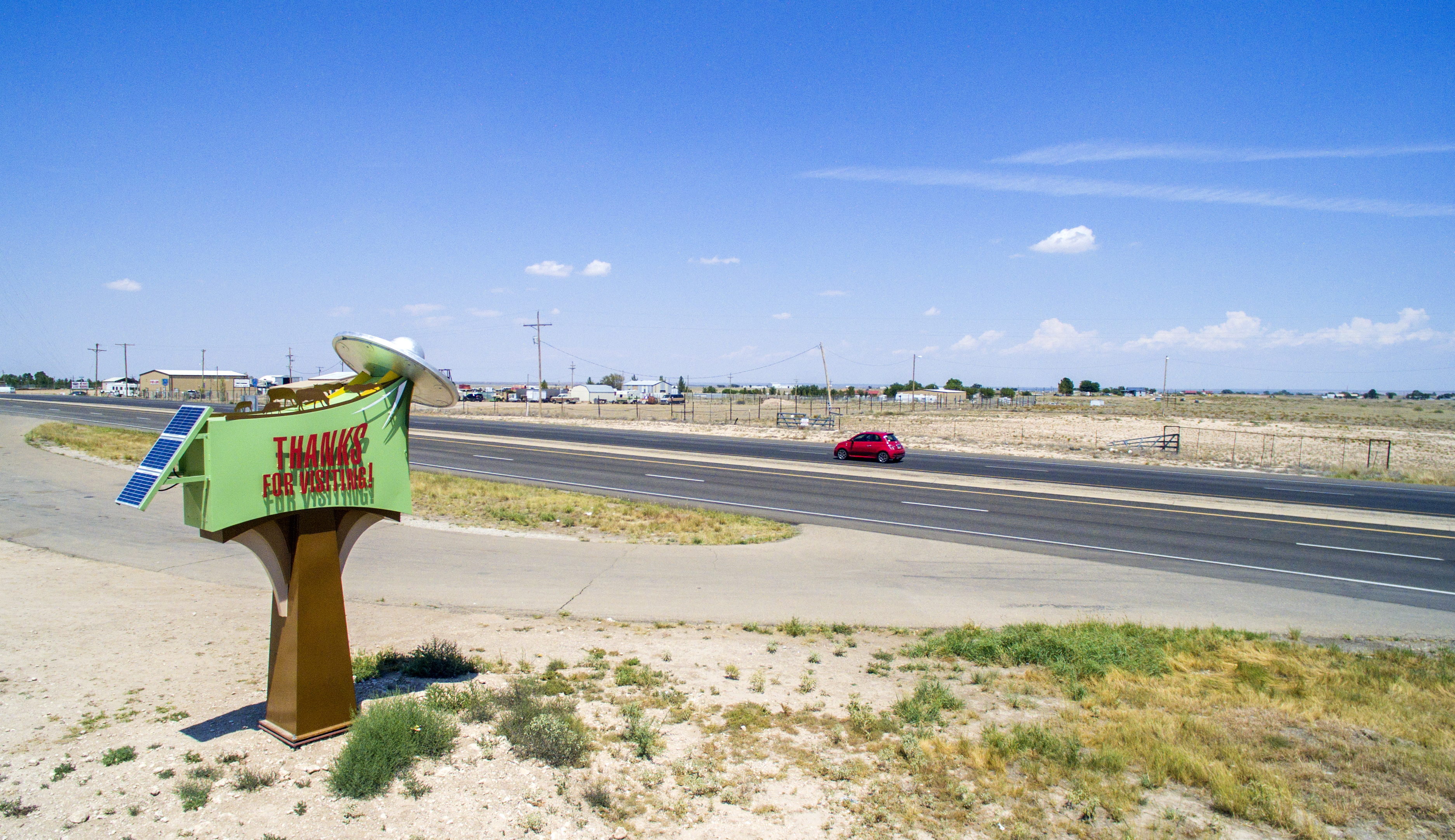 A colorful roadside sign labeled "THANK YOU FOR VISITING" beside a busy highway with a red car passing by and open fields under a clear blue sky.