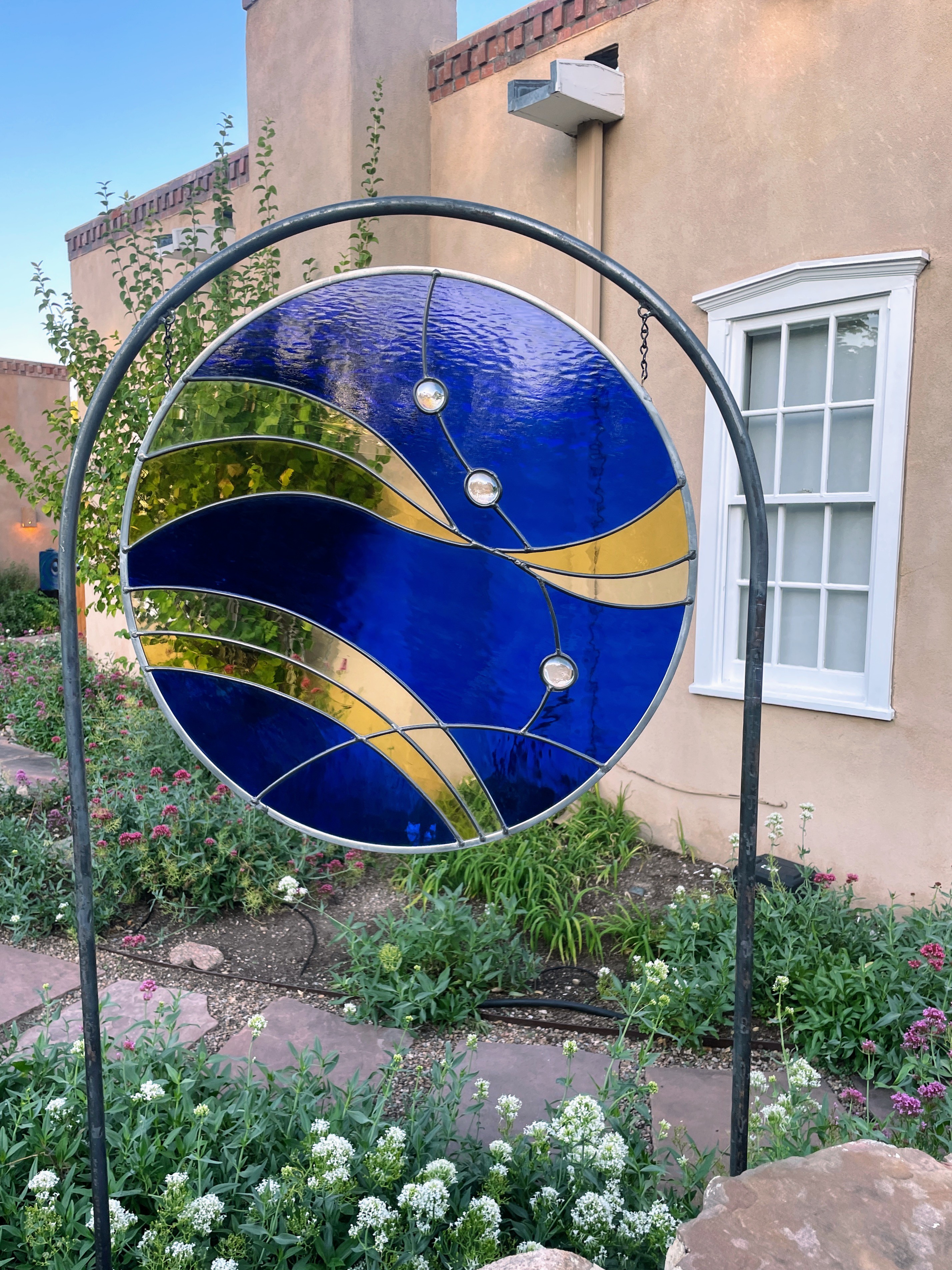 Decorative stained glass panel featuring blue and gold swirls with embedded clear crystals, set in a metal frame in front of a stucco building with a small window.