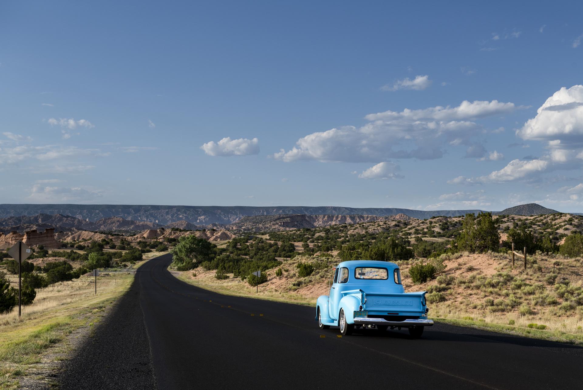 A vintage blue truck parked on the side of a winding road in a desert landscape with sparse vegetation and distant hills under a clear blue sky.