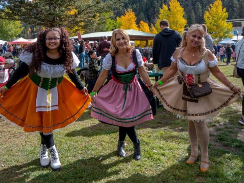 Three individuals in traditional Bavarian attire smiling at a festive outdoor event, surrounded by attendees, trees, and a mountainous backdrop.