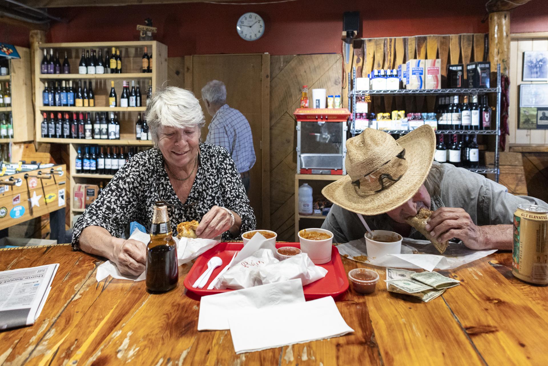 Two individuals enjoying a meal at a rustic restaurant with shelves filled with bottled products in the background. 