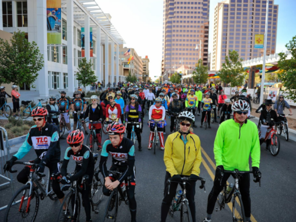 A group of cyclists gathered on a city street, ready to start a bike race. The setting includes modern buildings and clear skies.