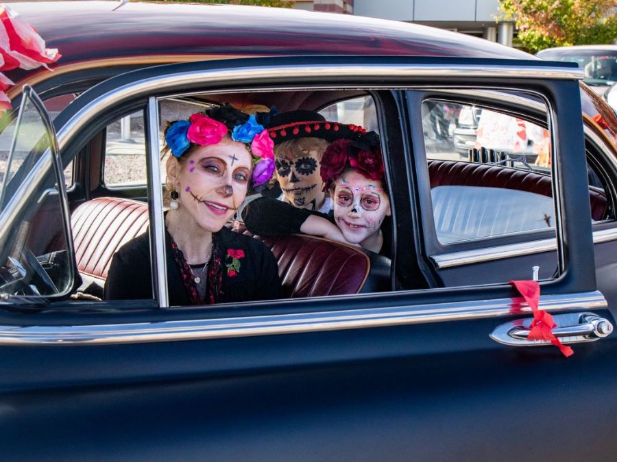 Three people sit in a classic car wearing face paint celebrating Dia de los Muertos