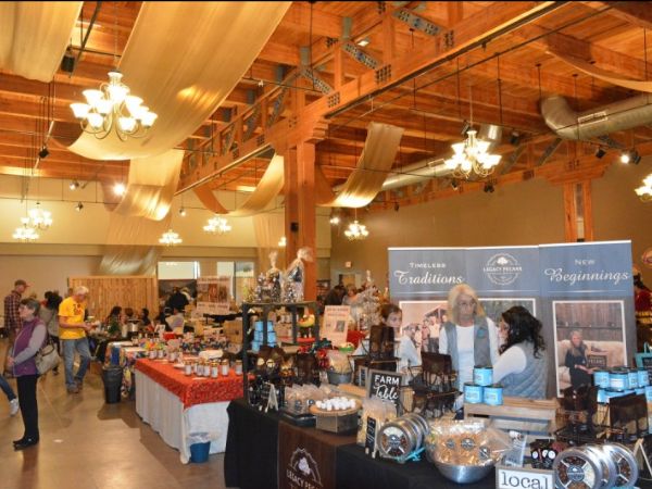 Interior of a bustling craft fair with various stalls displaying handmade goods and local products under warm lighting in a large hall with wooden beams.