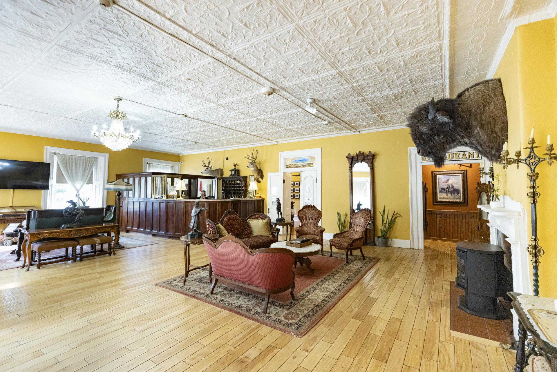 Interior view of a spacious room with vintage decor, featuring a wooden floor, elaborate ceiling patterns, and classic furniture. Notable decorations include a large stuffed animal head mounted above a fireplace and a chandelier hanging from the ceiling.