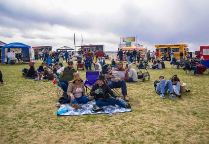 People sitting on grass at an outdoor festival with food trucks and tents in the background.