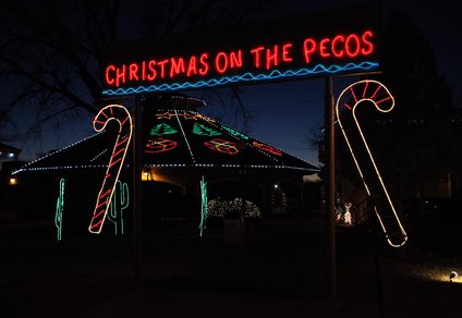 "Christmas on the Pecos" illuminated sign with festive lights including candy canes, stars, and Christmas tree designs displayed over a park structure at night.