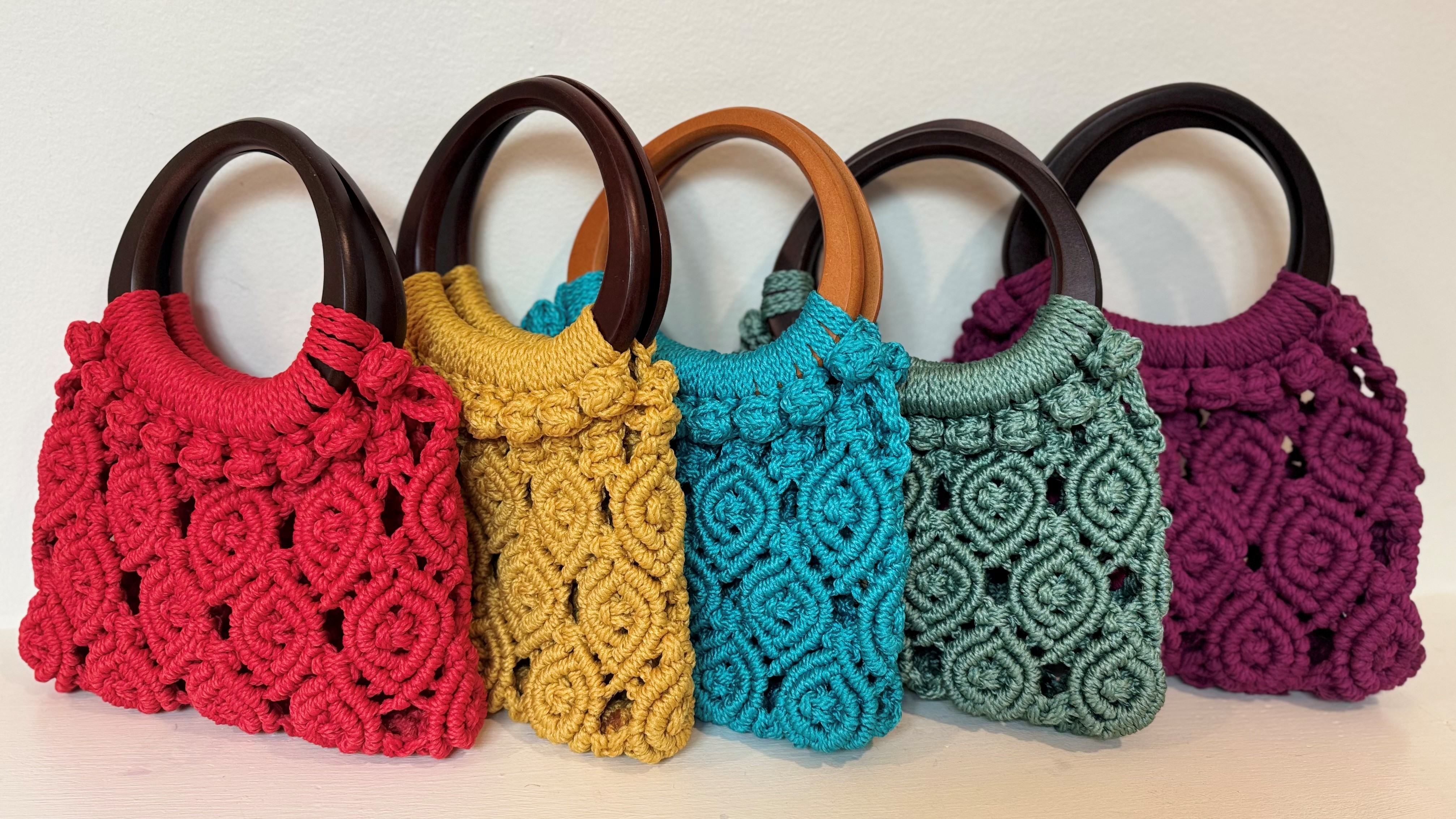 A row of five colorful macrame handbags, each featuring intricate patterns and a unique wooden handle, displayed against a white background.
