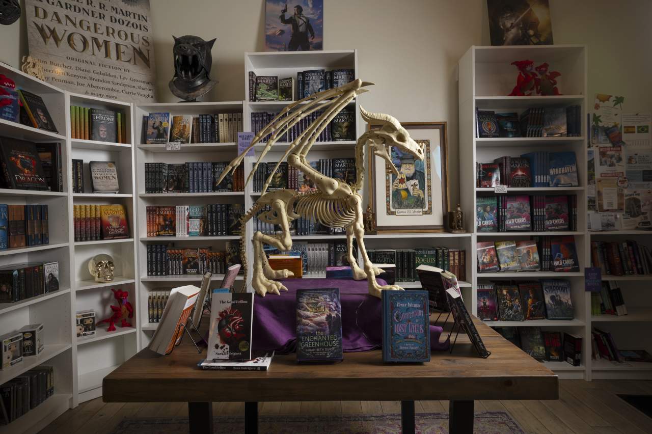 A skeleton of a mythical dragon is displayed prominently in a room filled with bookshelves stocked with numerous books. Featured in the foreground on a wooden table are several books.