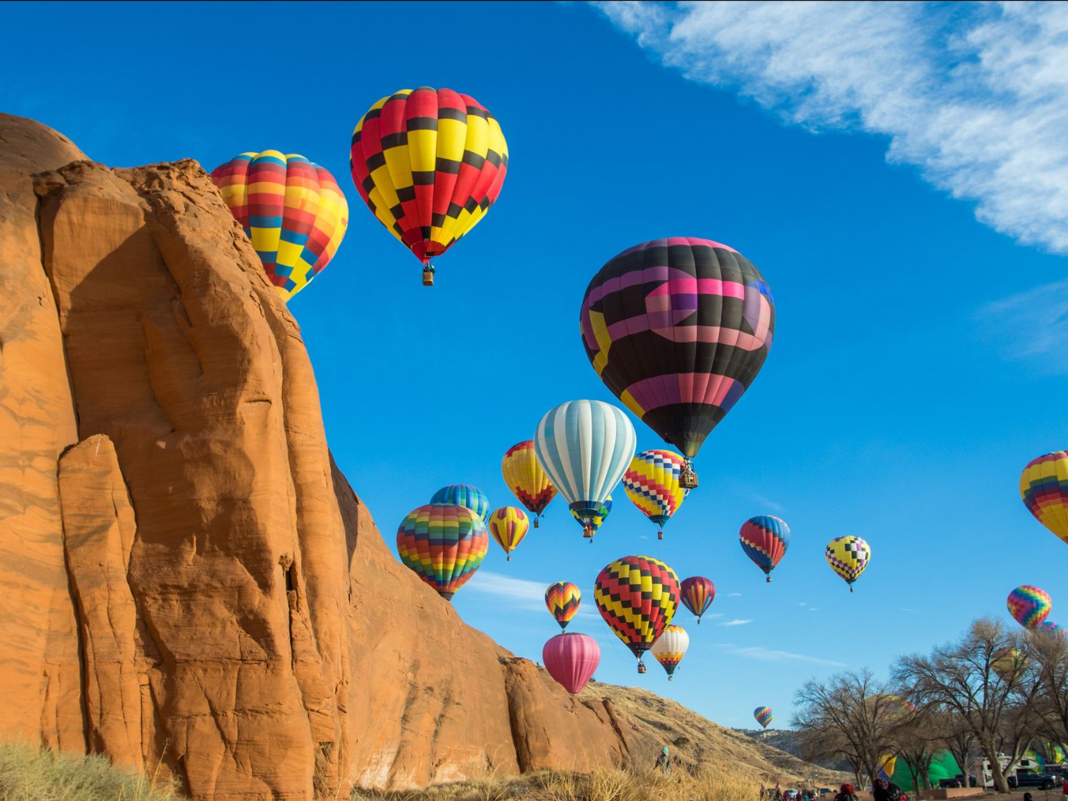 Colorful hot air balloons ascending in a clear blue sky near large red rock formations.